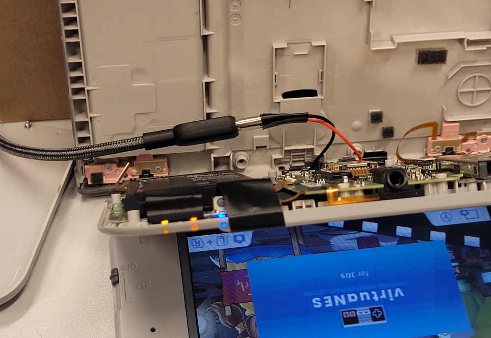 An upside-down New 3DS XL lying open on a desk. A small USB-C breakout board is attached to the 3DS, and a USB-C cable is plugged in. The 3DS is glowing to indicate that it is charging.
